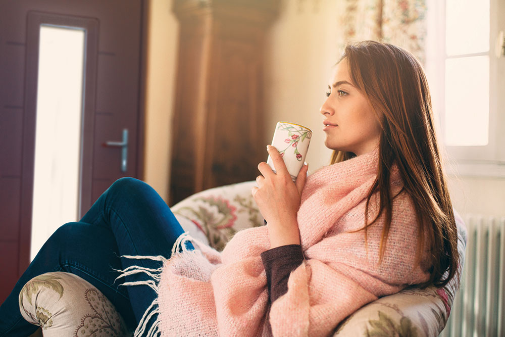 Woman relaxing on a sofa holding a mug wrapped in a pink blanket, with warm sunlight streaming through a composite door in the background