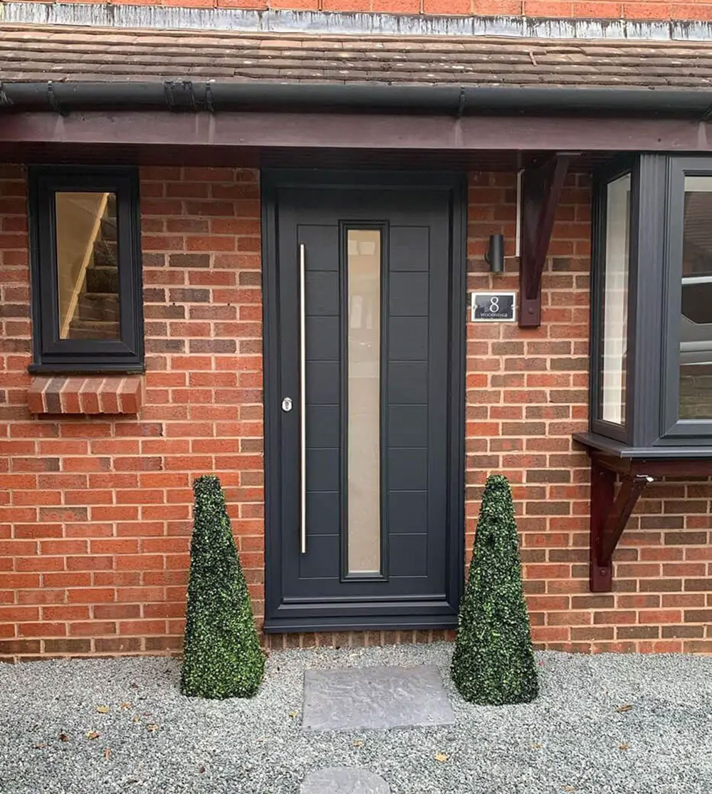 Anthracite grey composite front door with slim central glazed panel and brass bar handle, flanked by topiary cones on a red brick detached home